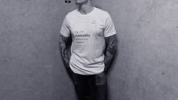 A man wearing an Assembly t-shirt leans against a concrete wall