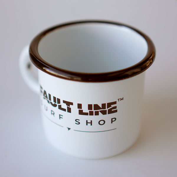 A three-quarter view of Fault Line's white enamel mug. The mug has a black lip. The front of the mug has a black Fault line logo on it.