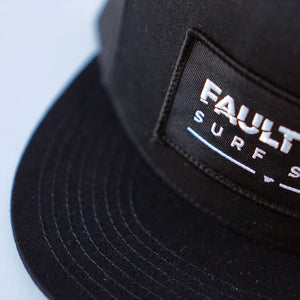 A close-up of Fault Line's black snapback trucker cap. The front of the cap has an embroidered patch with the logo on it.