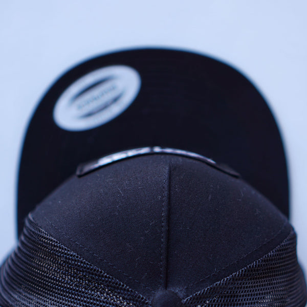 An overhead view of Fault Line's black snapback trucker cap. The front of the cap has an embroidered patch with the logo on it.