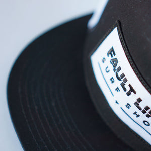 A close-up of Fault Line's black & white snapback trucker cap. The front of the cap has an embroidered patch with the logo on it.